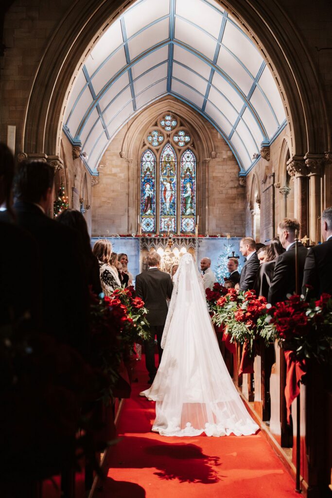 Bridal gown from behind as the bride walks down the aisle with here dad in the church in All Saints Church Essex